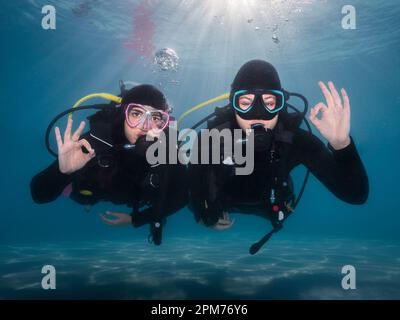 Closeup of two happy scuba divers underwater facing the camera showing the ok sign with the bright sunrays shining through the water surface behind th Stock Photo