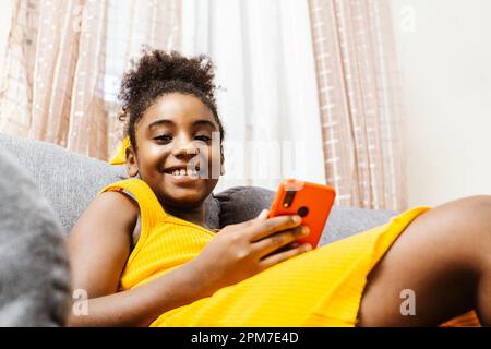 A cheerful African-American girl is sitting on a sofa while using a mobile device Stock Photo