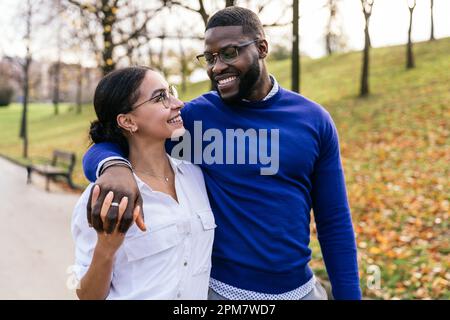 Interracial Couple Embracing in Autumn Park: Handsome Black Boy and Beautiful Caucasian Girl Walking and Smiling Happily at Sunset Stock Photo