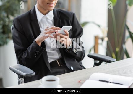 partial view of african american businesswoman in black blazer messaging on mobile phone near blurred coffee cup in office,stock image Stock Photo