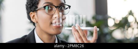 portrait of african american businesswoman in stylish eyeglasses sending voice message on smartphone in office, banner,stock image Stock Photo