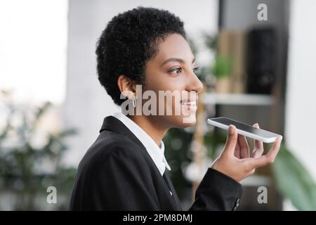 profile of positive african american businesswoman sending voice message on mobile phone while looking away in office,stock image Stock Photo