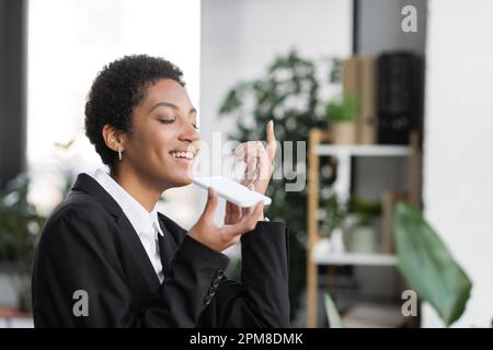 cheerful african american businesswoman holding smartphone with white screen while sending voice message in office,stock image Stock Photo