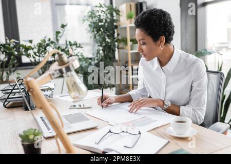 young african american businesswoman in white blouse writing on documents near laptop and coffe cup in modern office,stock image Stock Photo