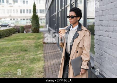 fashionable african american businesswoman in sunglasses standing with laptop and takeaway drink near city building with glass facade,stock image Stock Photo