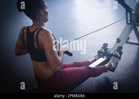 Rear view of biracial young woman exercising on rowing machines in gym, copy space Stock Photo
