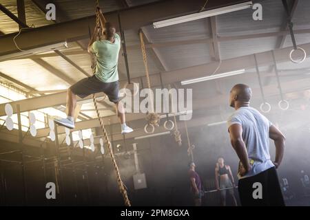 Biracial young man looking at male friend climbing on rope in gym, copy space Stock Photo