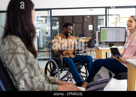 Diverse man sitting on wheelchair and using tablet while discussing with female coworkers in office Stock Photo