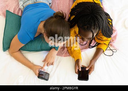 Happy diverse teenager girls friends lying on bed and using smartphones Stock Photo