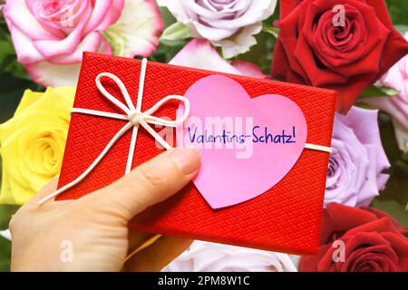FOTOMONTAGE, Hand hält Geschenk mit einem herzförmigen Zettel und der Aufschrift Valentins-Schatz vor einem Blumenstrauß