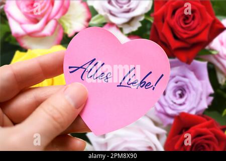 FOTOMONTAGE, Hand hält herzförmigen Zettel mit Aufschrift Alles Liebe vor einem Strauß Rosen