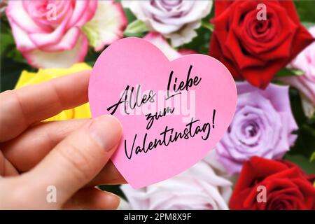 FOTOMONTAGE, Hand hält herzförmigen Zettel mit Aufschrift Alles Liebe zum Valentinstag vor einem Strauß Rosen