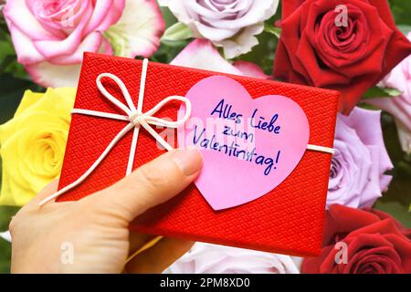 FOTOMONTAGE, Hand hält Geschenk mit einem herzförmigen Zettel und der Aufschrift alles Liebe zum Valentinstag vor einem Blumenstrauß