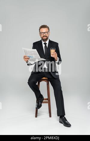 full length of happy businessman in suit and glasses holding newspaper while sitting with paper cup on grey,stock image Stock Photo