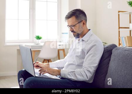 Mature man doing remote work working on laptop in his home office making notes sitting on couch. Stock Photo