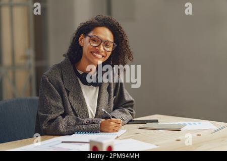 Pretty woman freelancer making notes in notebook at desk in modern coworking and looking at camera Stock Photo