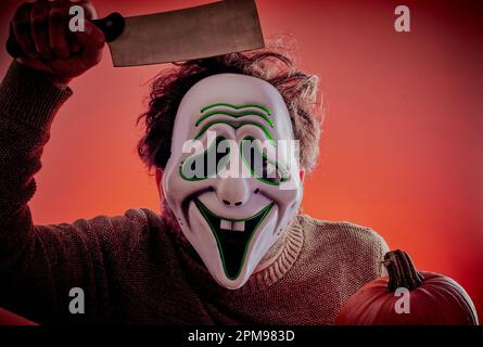 Man dressed up for Halloween with a scary mask holding a pumpkin with a big knife in his hand. Stock Photo