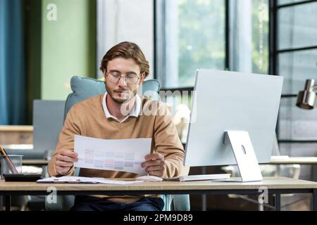 A young businessman, freelancer, programmer sits in the office at a table with a computer, works with documents, schedules, bills, on a project, deadline overtime.. Stock Photo