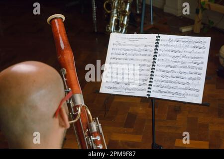 rear view of bald man sitting at home playing bassoon, reading sheet music at music stand, practicing and studying classical music. copy space. focus Stock Photo
