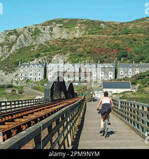 Woman cyclist completes a bike ride journey beside the historical Victorian Barmouth Bridge (or Pont Abermaw) or (Barmouth Viaduct) which is a Grade II listed single track wooden railway structure. Parallel to the train track is this footbridge walkway & both cross the estuary of the Afon Mawddach River linking Barmouth here here to Morfa Mawddach a small station 820 metres  away both in Gwynedd in North Wales. The footbridge is part of the National Cycle Route 8 & is available to pedestrians cyclists and motorbike users. Stock Photo