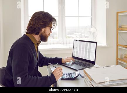 Male accountant calculates company's financial performance using calculator and laptop software. Stock Photo