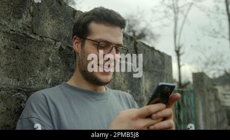 Handsome Young Man with Glasses Surprised and Smiling While Using Smartphone Outdoors Stock Photo