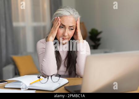 Despaired sad caucasian old lady sitting at table, looking at computer, suffering from headache, menopause, stress Stock Photo