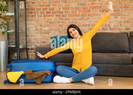 Cheerful mexican lady sitting near packed suitcase and making airplane gesture, feeling happy about abroad vacation Stock Photo