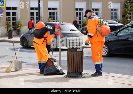 Two janitors in orange uniform sweeping the street on cars background. Man and woman workers cleaning spring city Stock Photo