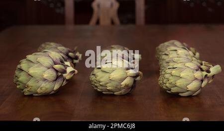 Several artichokes lie next to each other on a rustic wooden table. In the background an old wooden cupboard and a detail of a wooden chair. Stock Photo