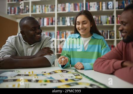 Diverse group of three people playing board game together with focus on smiling young woman throwing dice Stock Photo