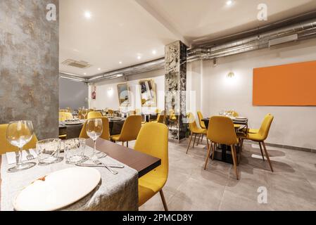 Dining room of a restaurant with tables, with cutlery, crockery, glassware and assembled chairs upholstered in yellow fabric and exposed air condition Stock Photo
