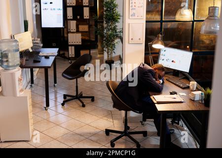 Entrepreneur doing overtime work in startup office, falling asleep desk trying to finish marketing strategy for company project. Employee working late at night, sleeping on table at workspace Stock Photo