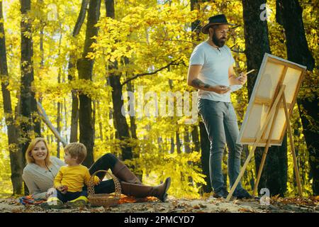 Handsome bearded man draws a picture of his young family in the park using a palette with paints and a spatula. Drawing easel and canvas with a Stock Photo