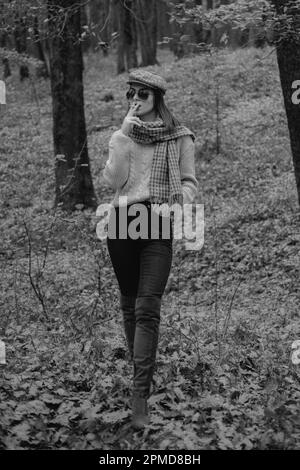 Autumn woman, Brunette girl is smoking a cigarette in the park during autumn. Fashionable model wearing warm brown boots and cap in the forest Stock Photo