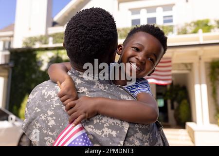 Happy african american boy with flag of america embracing army soldier father outside house Stock Photo