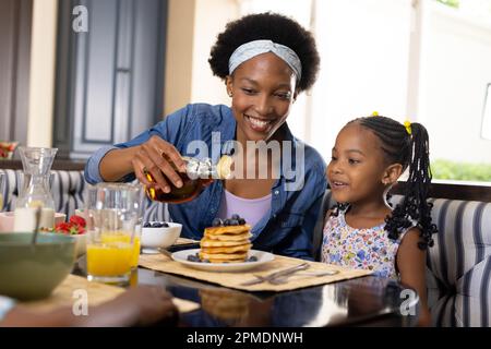 African american smiling mother pouring honey on pancakes while having breakfast with daughter Stock Photo
