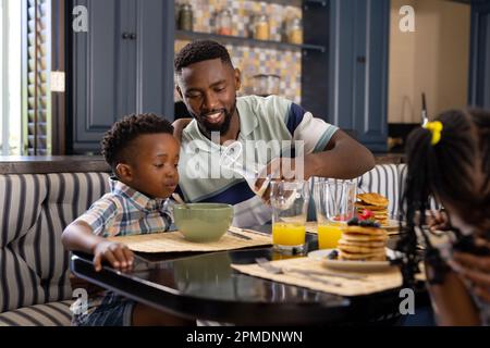 African american father pouring milk in bowl for son while having breakfast at dining table Stock Photo
