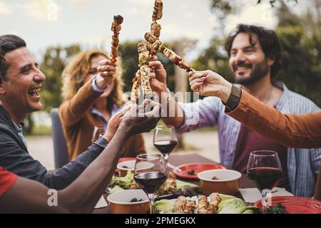 A group of people in their 30s and 40s sitting at a garden table, holding meat skewers and toasting. Focus on skewers. Mixed ethnicity, including a yo Stock Photo