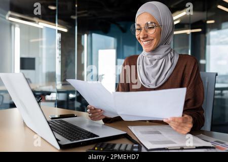 Successful muslim woman in hijab working inside office with documents, joyful financial accountant happy with reports checking financial data and smiling, businesswoman at workplace with laptop. Stock Photo