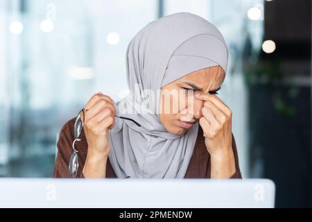 Tired and overworked business woman in hijab at workplace, muslim woman massaging eyes with glasses off, female worker working late on project. Stock Photo