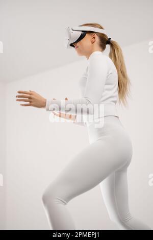 Low angle view of beautiful young futuristic looking woman wearing VR technology exploring the virtual reality in metaverse. Clean contemporary and mi Stock Photo