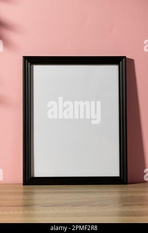 Vertical of black empty frame with copy space on table against pink wall and shadow of leaves Stock Photo