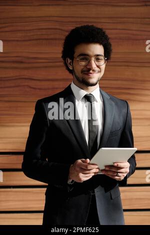 Portrait of young businessman in black suit smiling at camera while using digital tablet standing against wooden wall Stock Photo