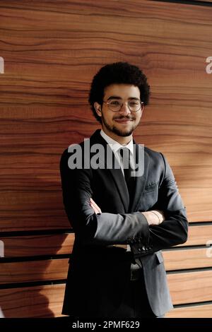 Portrait of young confident businessman in black suit standing with his arms crossed against wooden wall and looking at camera Stock Photo