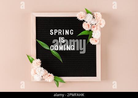 Letter board with Spring Is Coming text and flowers on beige wall. Blooming floral wooden frame Stock Photo