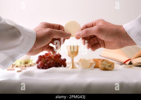 Hands of a priest consecrating a host as the body of Christ to distribute it to the communicants with table with sacred objects behind. Stock Photo