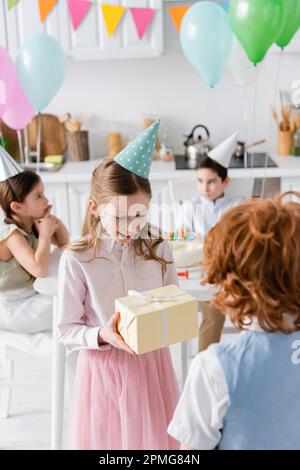 amazed girl in party cap receiving birthday present from redhead boy,stock image Stock Photo