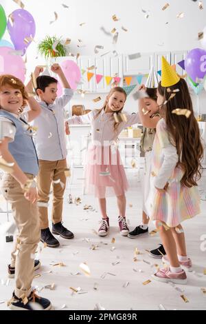 happy kids in party caps dancing under falling confetti during birthday celebration at home,stock image Stock Photo