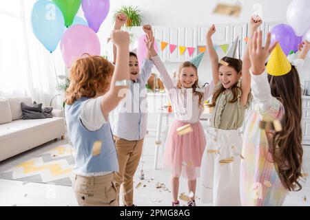 cheerful kids in party caps dancing under falling confetti during birthday celebration at home,stock image Stock Photo